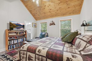 A bedroom with a wooden ceiling, a large bed with a quilt, a bookshelf, a painting, and a window with blinds.