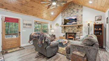 A cozy living room with a stone fireplace, wooden ceiling, and a large window overlooking a forest.