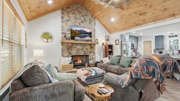 A cozy living room with a stone fireplace, comfortable seating, and a kitchen in the background.