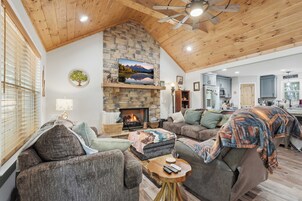 A cozy living room with a stone fireplace, comfortable seating, and a kitchen in the background.