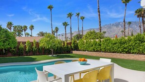 Un espace aménagé au bord de la piscine, avec une table et des chaises, un saladier rempli de fruits, et une vue sur les palmiers et les montagnes.