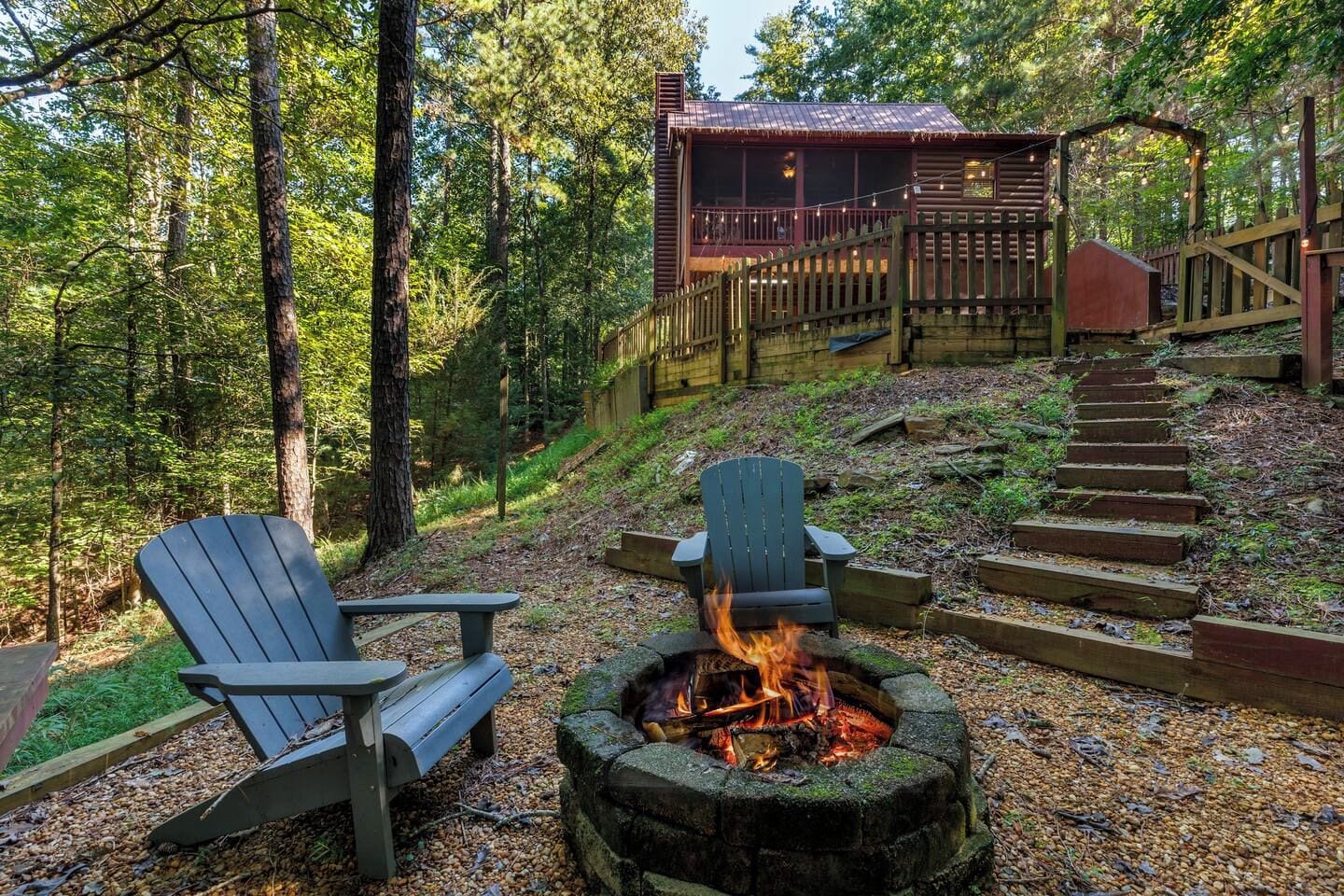 Metsäinen alue, jossa on nuotiopaikka, Adirondack-tyyliset puutarhatuolit ja puinen terassi, jolle johtaa portaat.