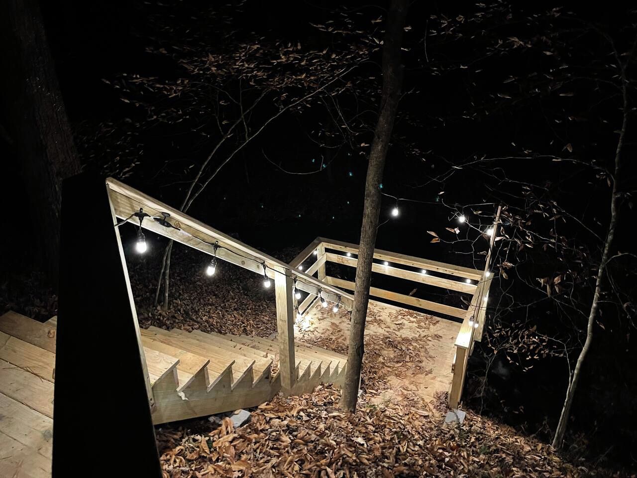 Un escalier en bois illuminé de guirlandes lumineuses, entouré d’arbres et de feuilles mortes.