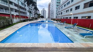 A swimming pool with clear blue water, surrounded by lounge chairs and a wooden deck. - SHB - Alto Padrão com Lazer Completo (Guarujá)
