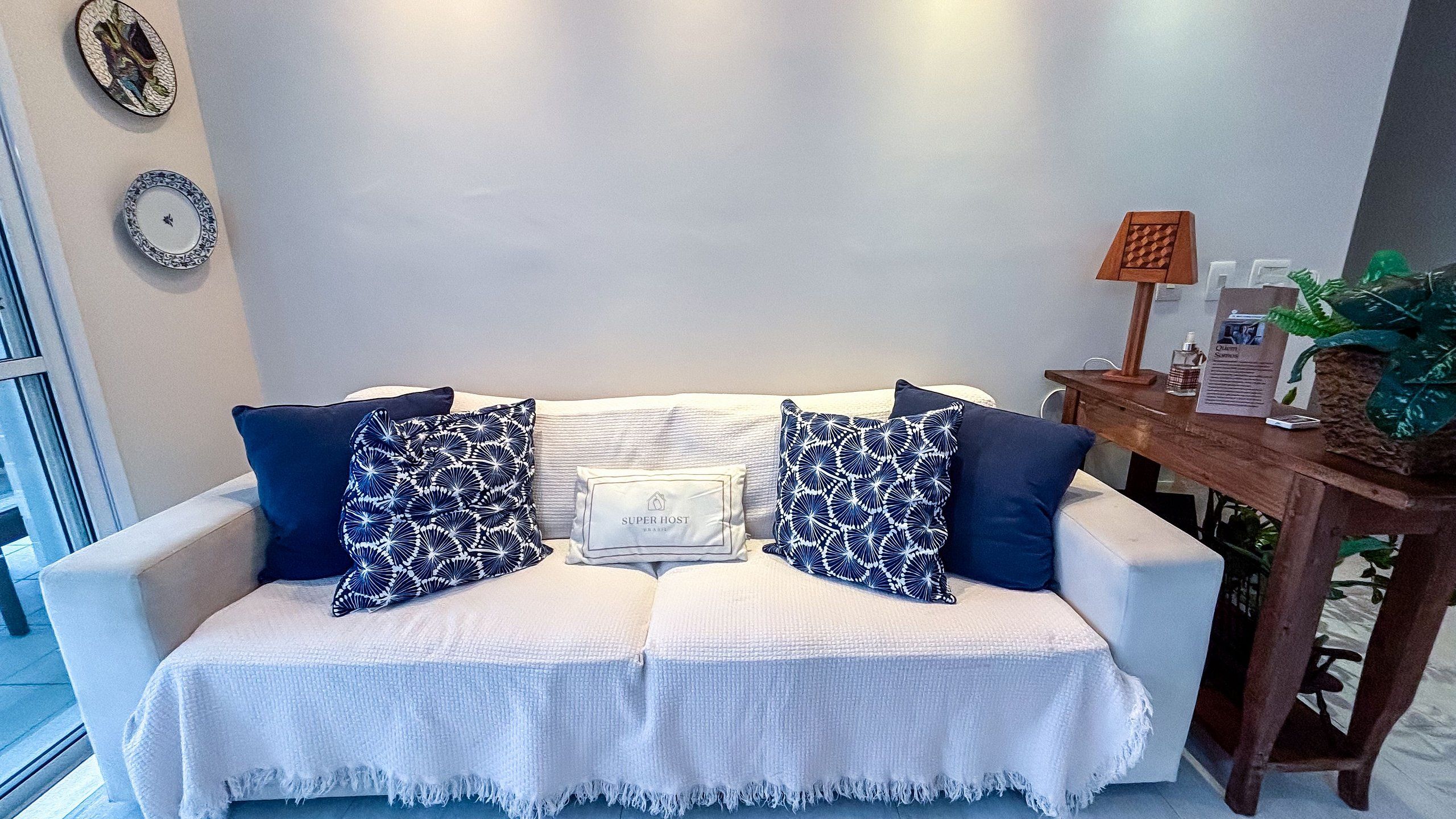 Un sofá blanco con dos almohadones estampados azules, una manta blanca y una mesita de madera con una planta y una botella.