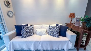 Un sofá blanco con dos almohadones estampados azules, una manta blanca y una mesita de madera con una planta y una botella.