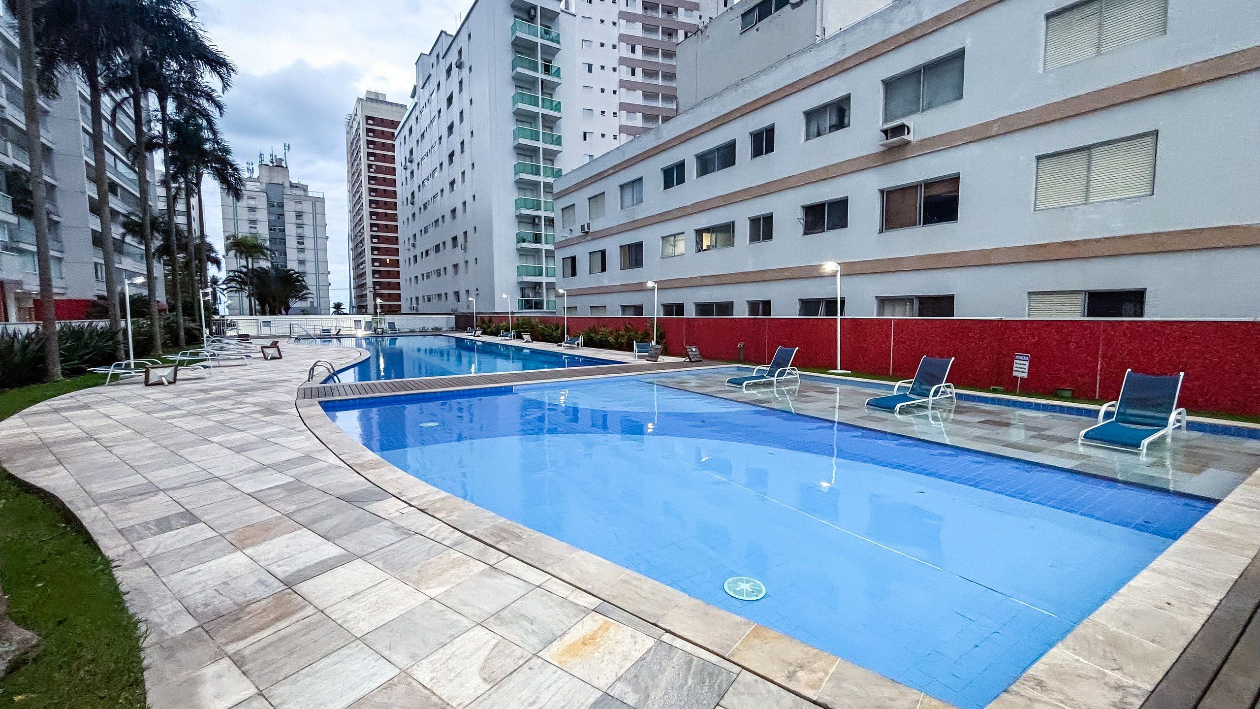 Una piscina con sillas de descanso, rodeada por un muro rojo y un edificio de varios pisos.