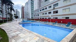 A swimming pool with lounge chairs, surrounded by a red wall and a building with multiple floors. - SHB - Alto Padrão com Lazer Completo (Guarujá)