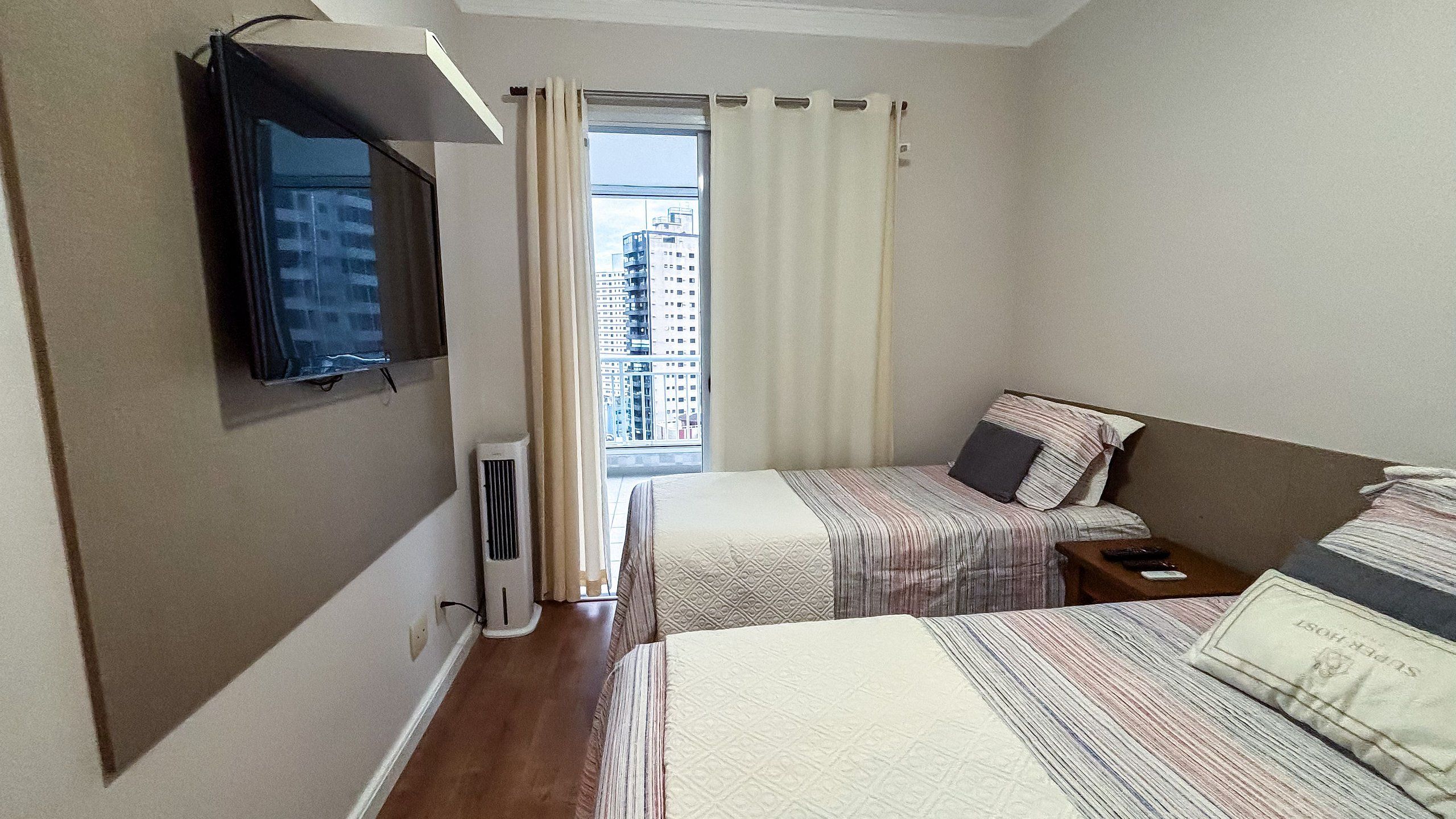 Quarto de hotel com duas camas, uma televisão presa à parede, janela com cortinas e vista para prédios lá fora.