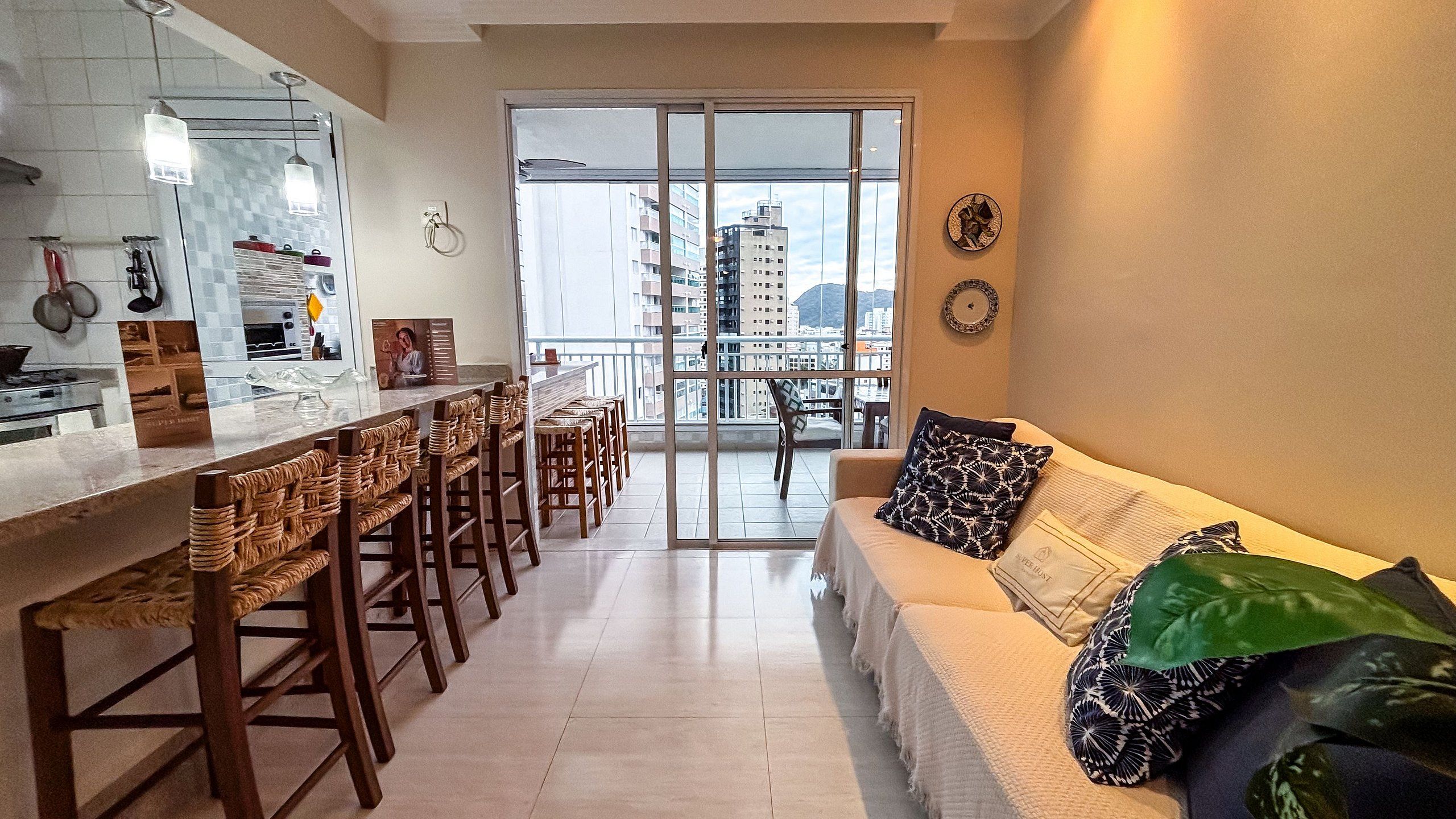 Cozinha moderna com área de jantar, varanda com vista da cidade e sala de estar com sofá e almofadas.