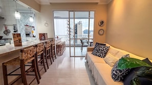 A modern kitchen with a dining area, a balcony with a city view, and a living room with a sofa and cushions. - SHB - Alto Padrão com Lazer Completo (Guarujá)