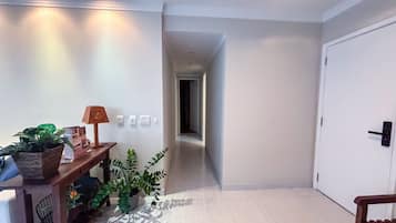 Um corredor com uma escrivaninha de madeira, uma luminária e plantas em vasos. Há interruptores de luz na parede e uma porta no final.