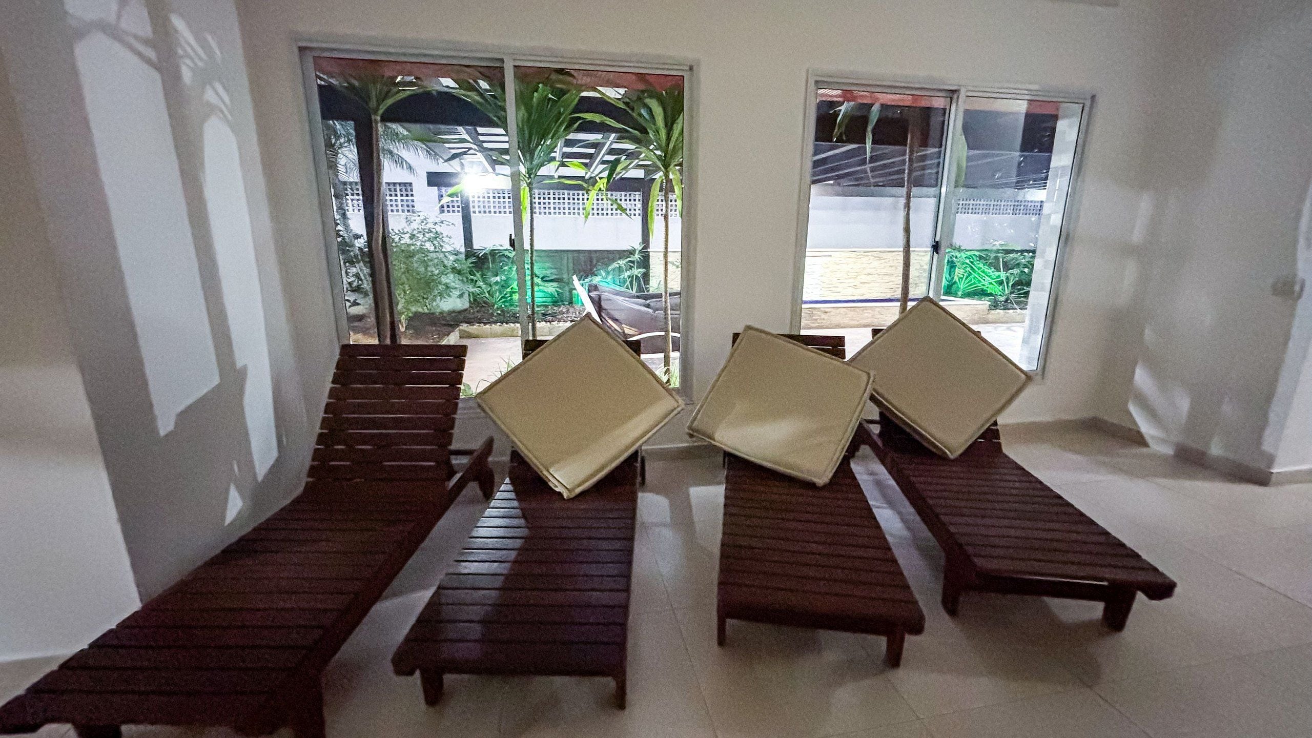 A room with four wooden lounge chairs and a small table, large windows with a view of greenery and a building, and a tiled floor.