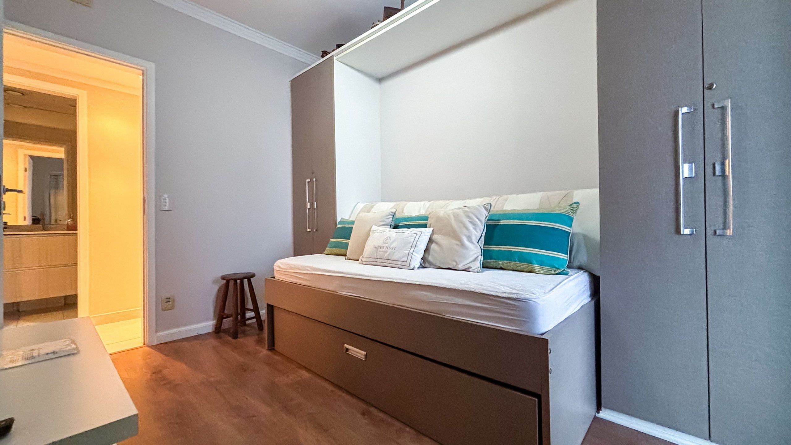 Un dormitorio compacto con cama, un taburete pequeño, un armario y una zona de kitchenette.