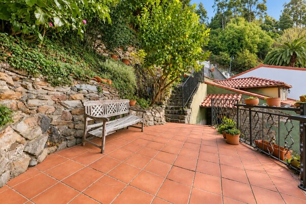 A stone wall with a bench, a tiled patio, and a house with a red-tiled roof.