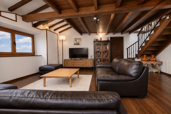 A modern living room with a wooden ceiling, leather sofas, a wooden coffee table, a TV, and a staircase.