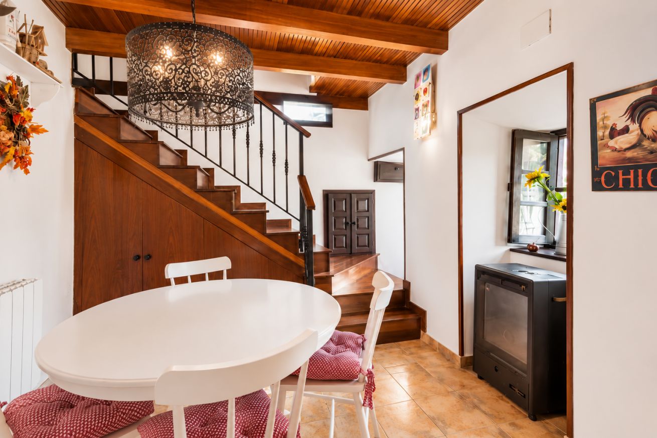 Un comedor con una mesa y sillas blancas, una escalera de madera y una zona de cocina con estufa.