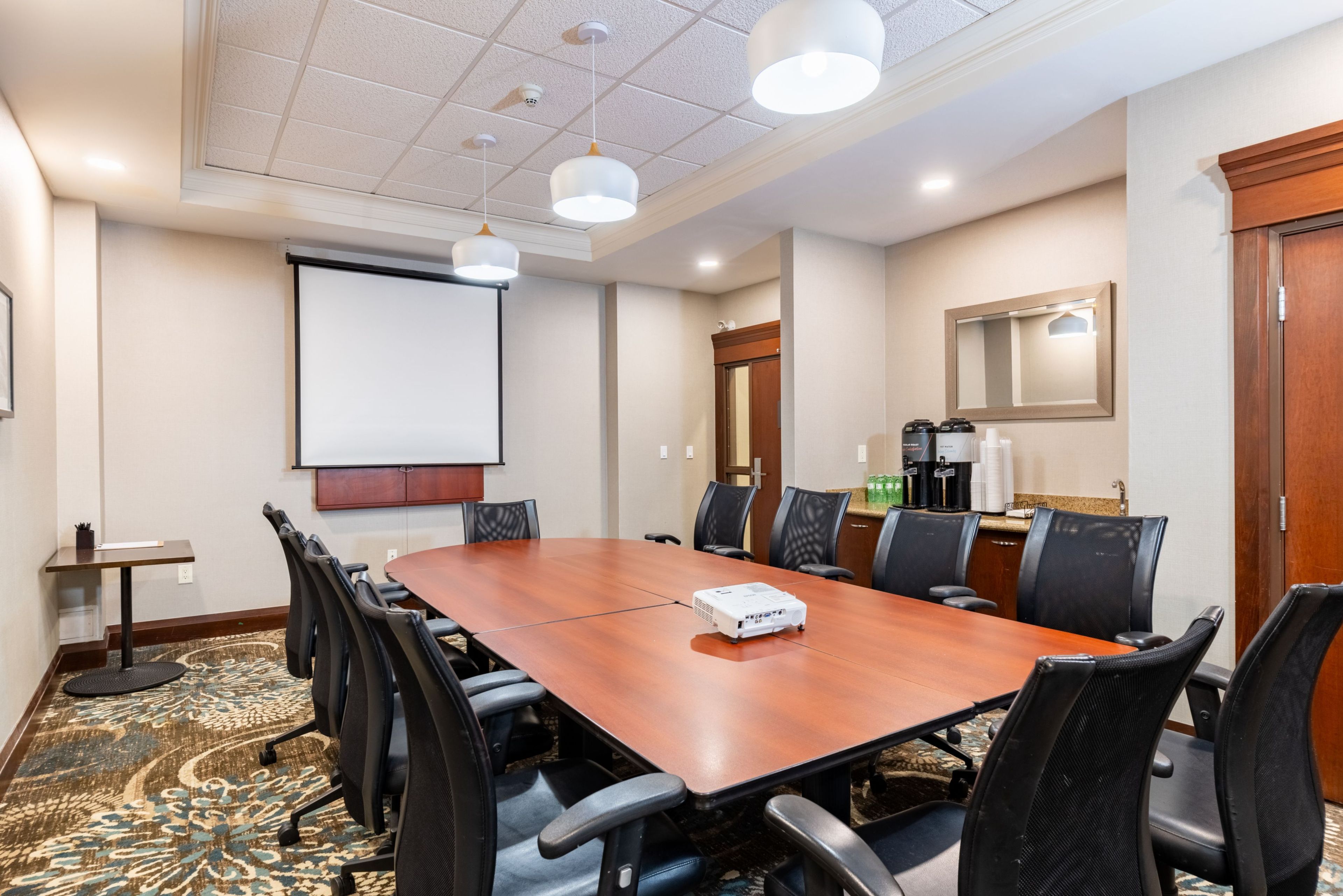A conference room with a large wooden table, black chairs, a projector, a screen, and a coffee station.