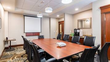 A conference room with a large wooden table, black chairs, a projector, a screen, and a coffee station.
