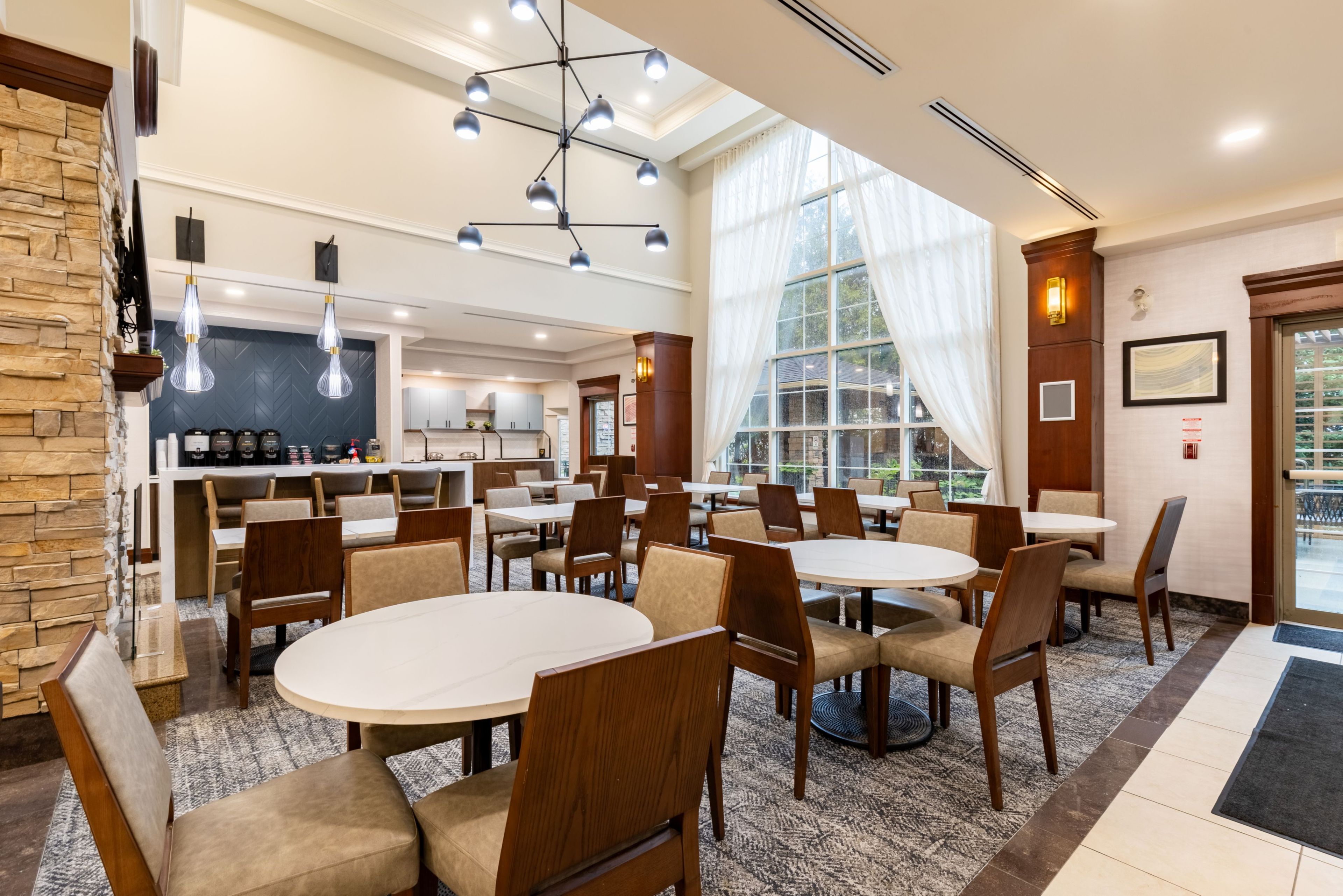A hotel dining area with round tables and chairs, a kitchen in the background, and large windows with curtains.
