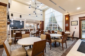 A hotel dining area with round tables and chairs, a kitchen in the background, and large windows with curtains.