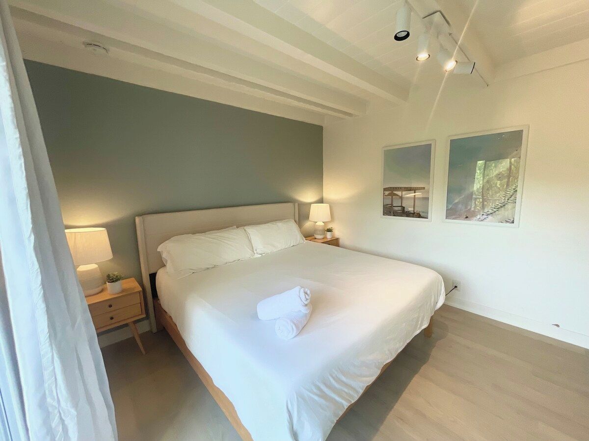A neatly made bed with white linens, a bedside lamp, and a small bedside table with a plant.