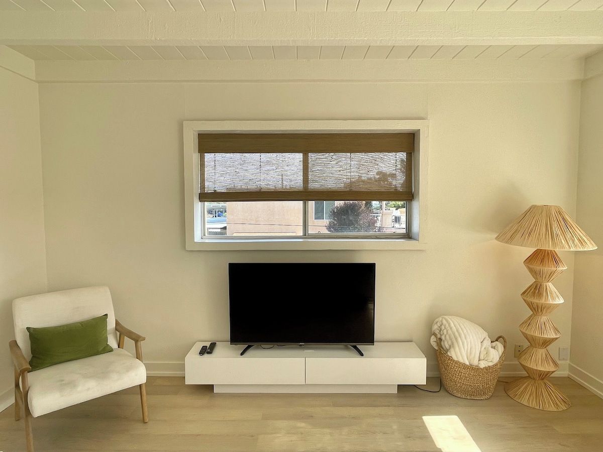 En stue med et fladskærms-tv på et hvidt skab, et vindue med udsigt, en lampe og en lænestol.