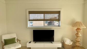En stue med et fladskærms-tv på et hvidt skab, et vindue med udsigt, en lampe og en lænestol.