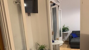 Ein Zimmer mit einem weißen Schminktisch, einem Spiegel, einem kleinen Tisch mit Topfpflanzen, einem grauen Sofa und einem Fliesenboden.