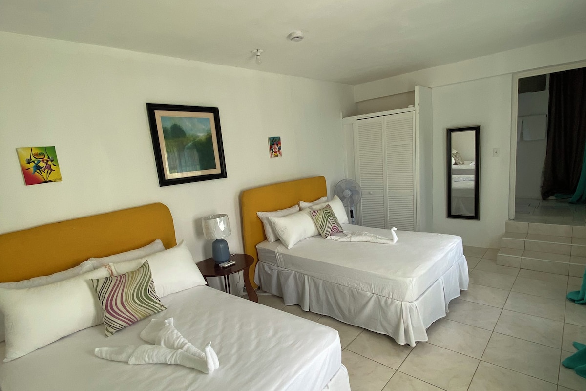 A hotel room with two beds, a bedside table with a lamp, a fan, and a door leading to a balcony.