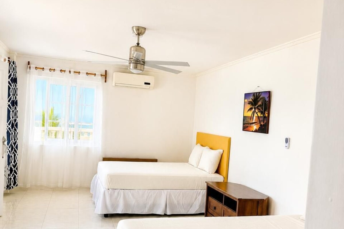 Una habitación de hotel con dos camas, ventilador de techo, equipo de aire acondicionado, ventana con cortinas y un cuadro en la pared.