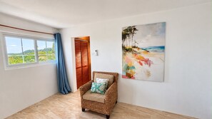 Una habitación con un ventanal, una silla de mimbre y un cuadro en la pared.