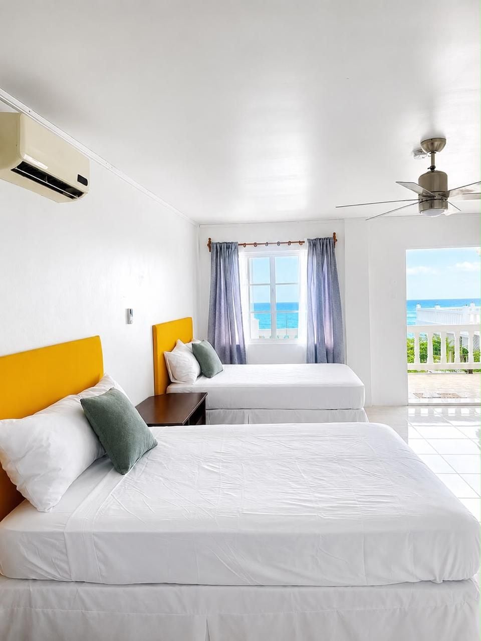 Una camera d'albergo con due letti, una ventola a soffitto, un condizionatore d'aria, una finestra con tende e vista sul mare.