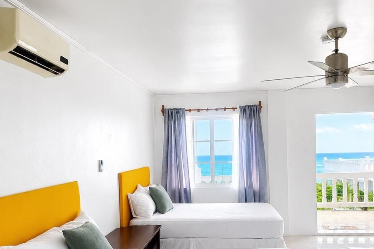 Habitación de hotel con dos camas, ventilador de techo, aire acondicionado, ventana con cortinas y vista al mar.