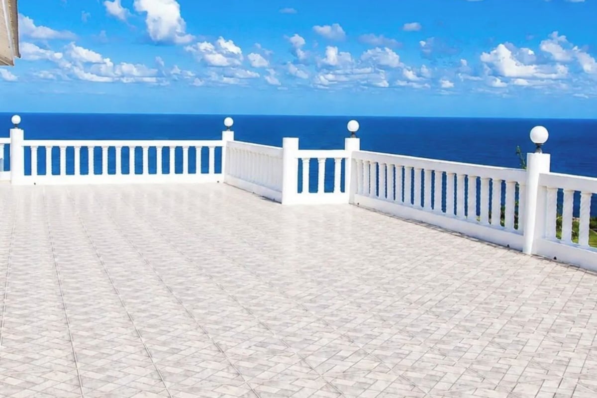 A white balcony with a tiled floor and a white railing, overlooking a clear blue sky and ocean.