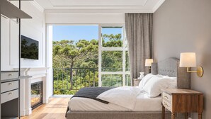 Ein Schlafzimmer mit Bett, Nachttischlampen, einem Fernseher, einem Kamin und einem Balkon mit Blick auf Bäume.