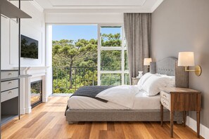 Ein Schlafzimmer mit Bett, Nachttischlampen, einem Fernseher, einem Kamin und einem Balkon mit Blick auf Bäume.