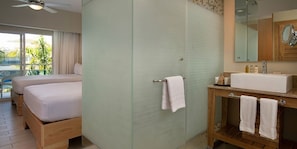 Ein Hotelzimmer mit Bett, einem Badezimmer mit Waschbecken und Spiegel sowie Blick auf Palmen durch das Fenster.