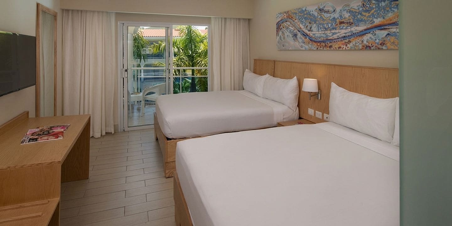 Habitación de hotel con dos camas, un escritorio, televisión y balcón con vista a palmeras.