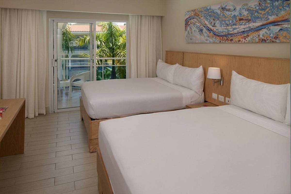 Ein Hotelzimmer mit zwei Betten, einem Schreibtisch, einem Fernseher und einem Balkon mit Blick auf Palmen.