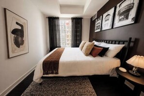 Un cuarto de hotel con cama, mesita de noche, lámpara, ventana con cortinas e imágenes enmarcadas en la pared.