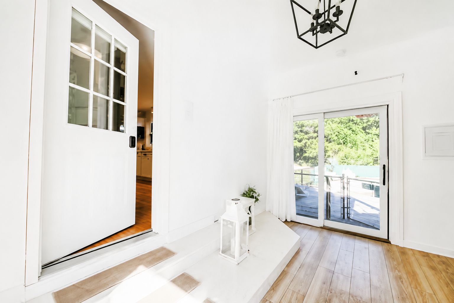 Un interior luminoso y blanco con piso de madera, una cocina visible a través de una puerta abierta y un cerramiento de vidrio corredero que da a una terraza exterior.