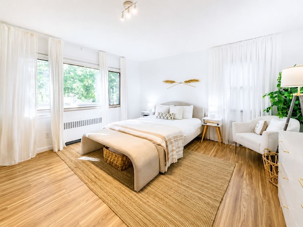 Une chambre d’hôtel moderne avec un grand lit, une table de chevet, une chaise, une lampe sur pied et une vue sur la verdure.