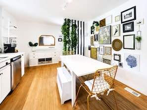 Une cuisine moderne avec des meubles blancs, un lave-vaisselle encastré et un coin repas comprenant une table blanche et une chaise en osier.