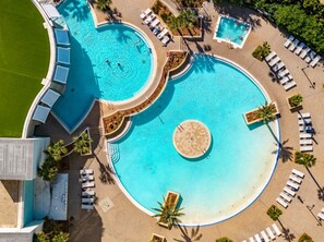 Une grande piscine circulaire, bordée d’une plage de sable, entourée de chaises longues et de palmiers.