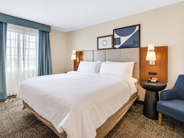 A hotel room with a large bed, two bedside tables with lamps, a chair, and a view of the outdoors through the window.