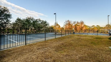 An outdoor basketball court with a metal fence, surrounded by trees and grass.