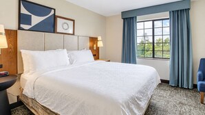 Quarto de hotel com uma cama grande, mesa de cabeceira, relógio de parede, janela com cortinas e vista para o verde lá fora.
