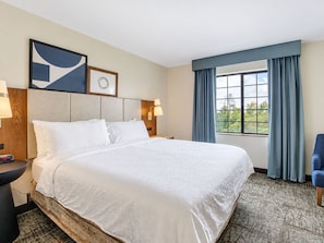Ein Hotelzimmer mit einem großen Bett, einem Nachttisch, einer Wanduhr, einem Fenster mit Vorhängen und Blick auf Grünflächen.