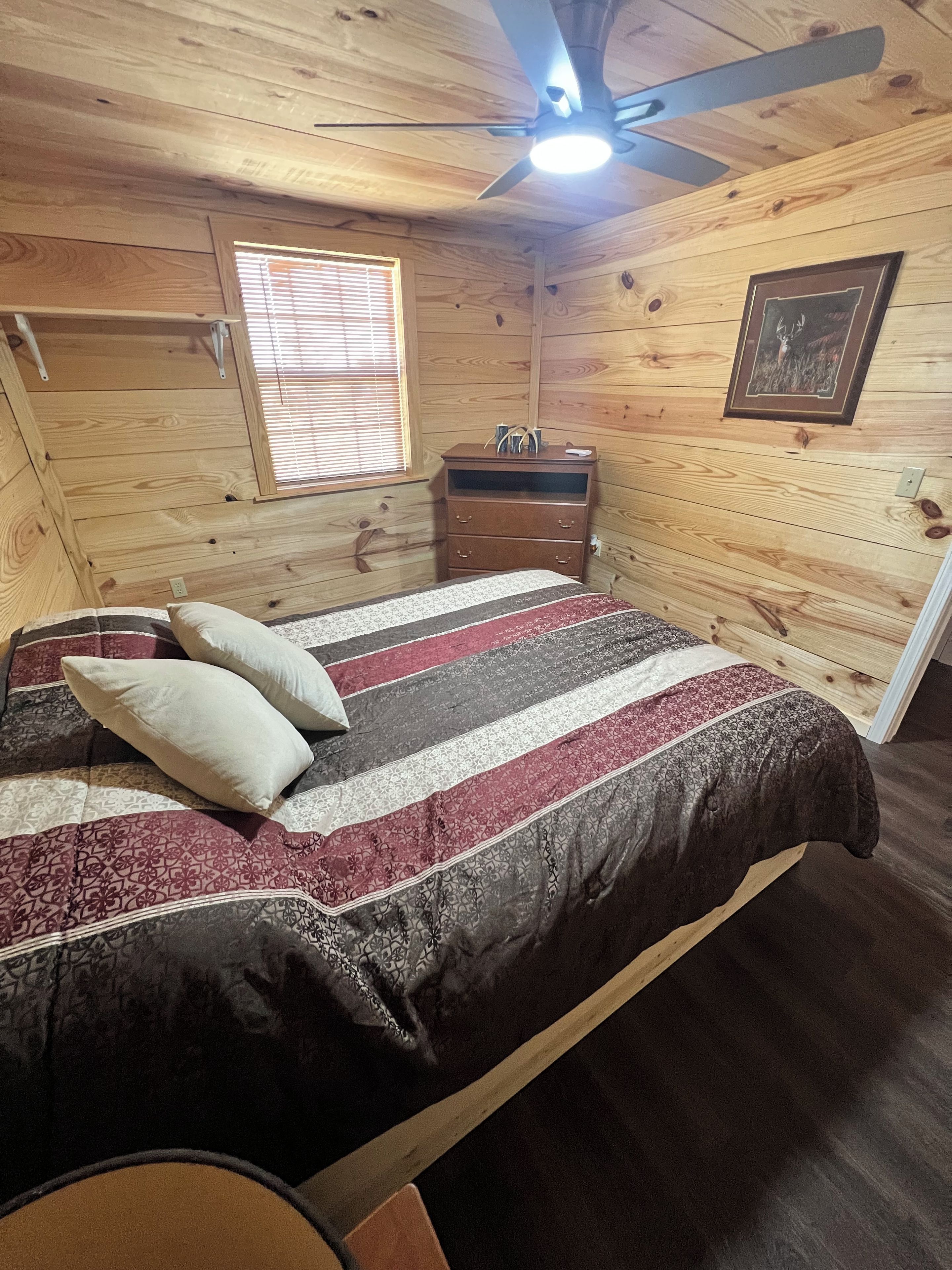 A wooden-paneled room with a bed, a ceiling fan, a window with blinds, a dresser, and a framed picture on the wall.